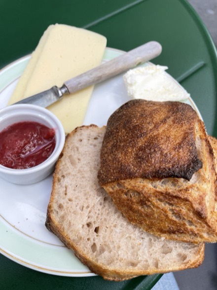 A plate of sliced sourdough with a crusty cheese bun, a wedge of cheese, butter and a ramekin of jam, on a green cafe table
